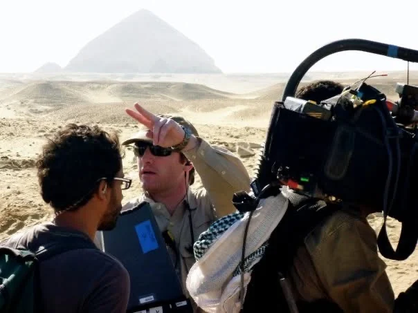 The Chasing Mummies crew working with a camera in the Egyptian desert, with a pyramid behind them.