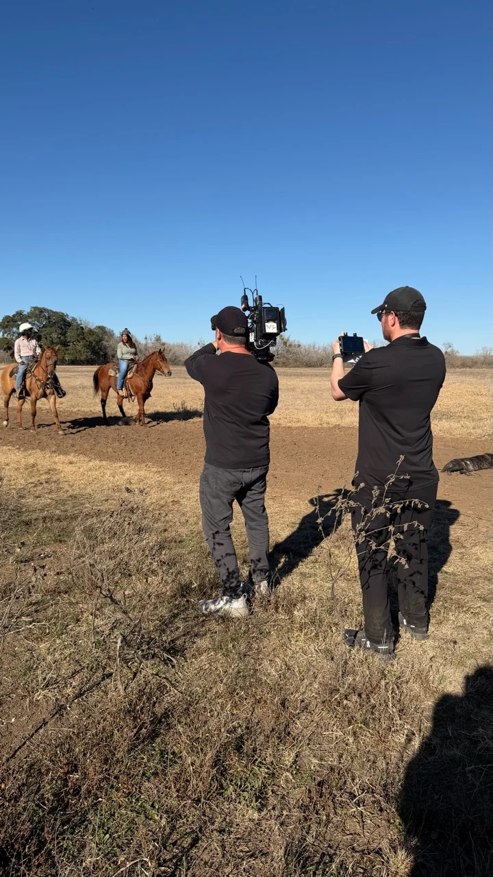 A camera crew films two riders on horseback in an open field.